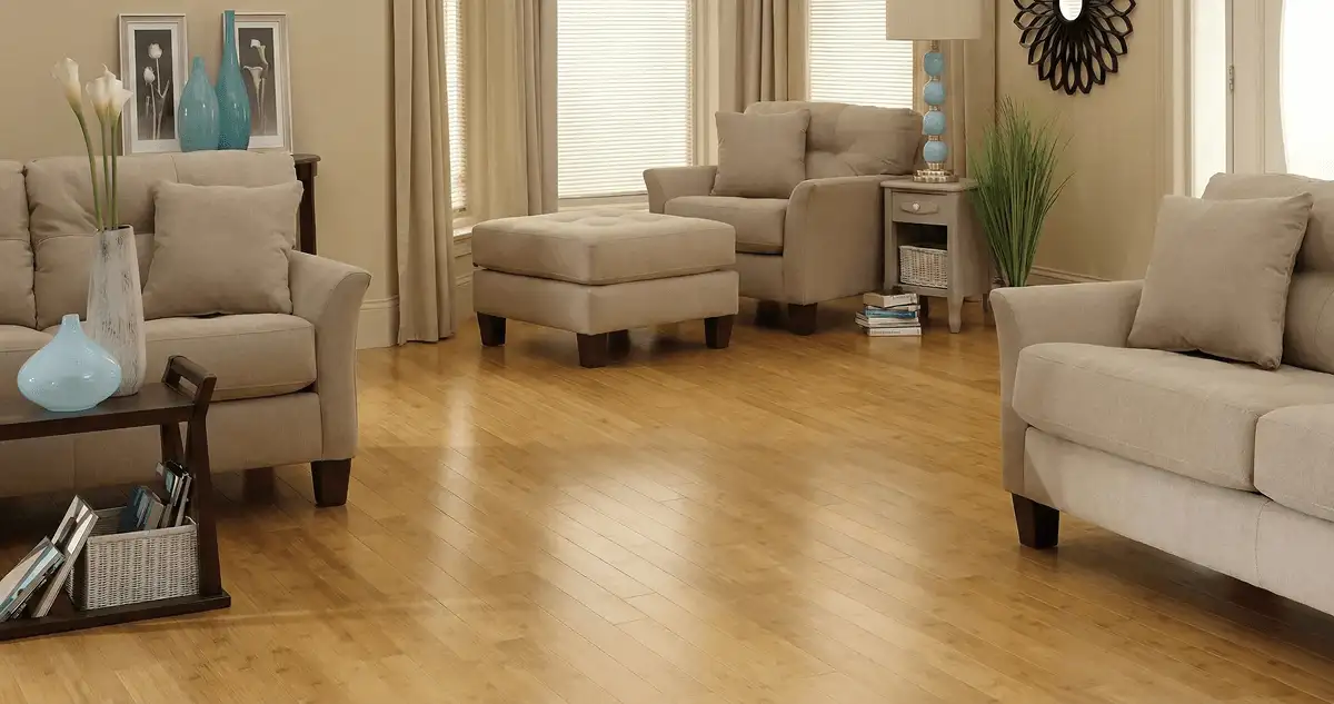 A living room with professionally installed bamboo flooring, beige sofas, a matching ottoman, a side table with a blue lamp, vases, framed pictures, and large windows with blinds and curtains in Tulsa, OK