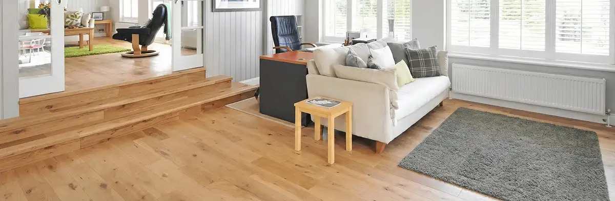 A modern living room with professionally installed laminate flooring, a beige sofa, a small side table, a gray rug, and large windows with white shutters in Tulsa, OK